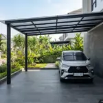 Carport 1 mobil lantai batu andesit 30×60 cm alur, kolom besi hollow hitam matte, atap polycarbonate UV, desain minimalis